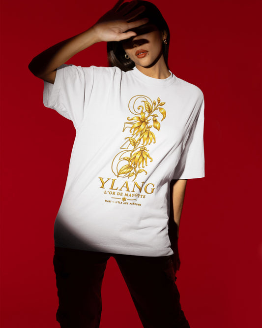 Femme portant le t-shirt oversize blanc YLANG — L’Or de Mayotte, avec fleurs dorées et inscription WASI, sur fond rouge.