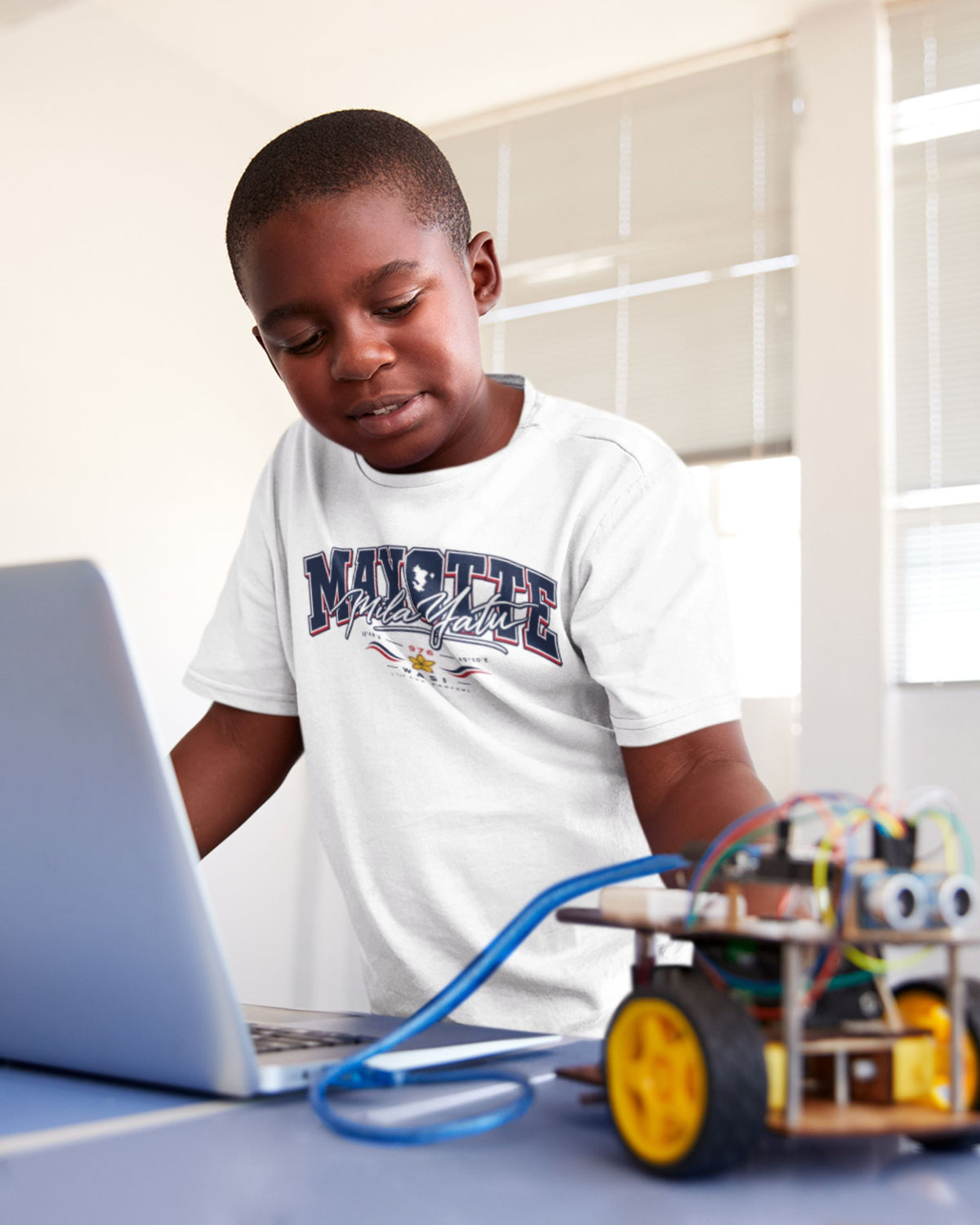Enfant portant le t-shirt blanc Mayotte Mila Yatu, regardant un ordinateur portable à côté d’un petit robot à roues.