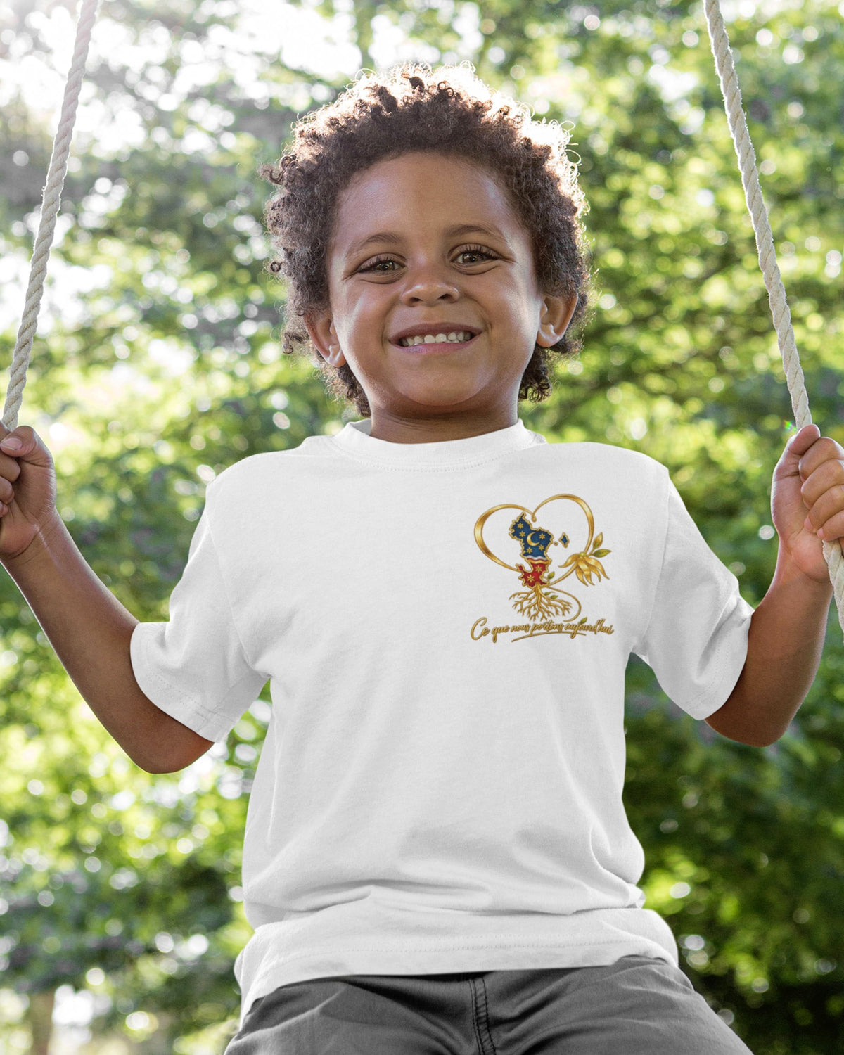 Enfant portant le t-shirt blanc WASI, avec cœur doré, carte de Mayotte et fleur d’ylang-ylang sur la poitrine, assis sur une balançoire.