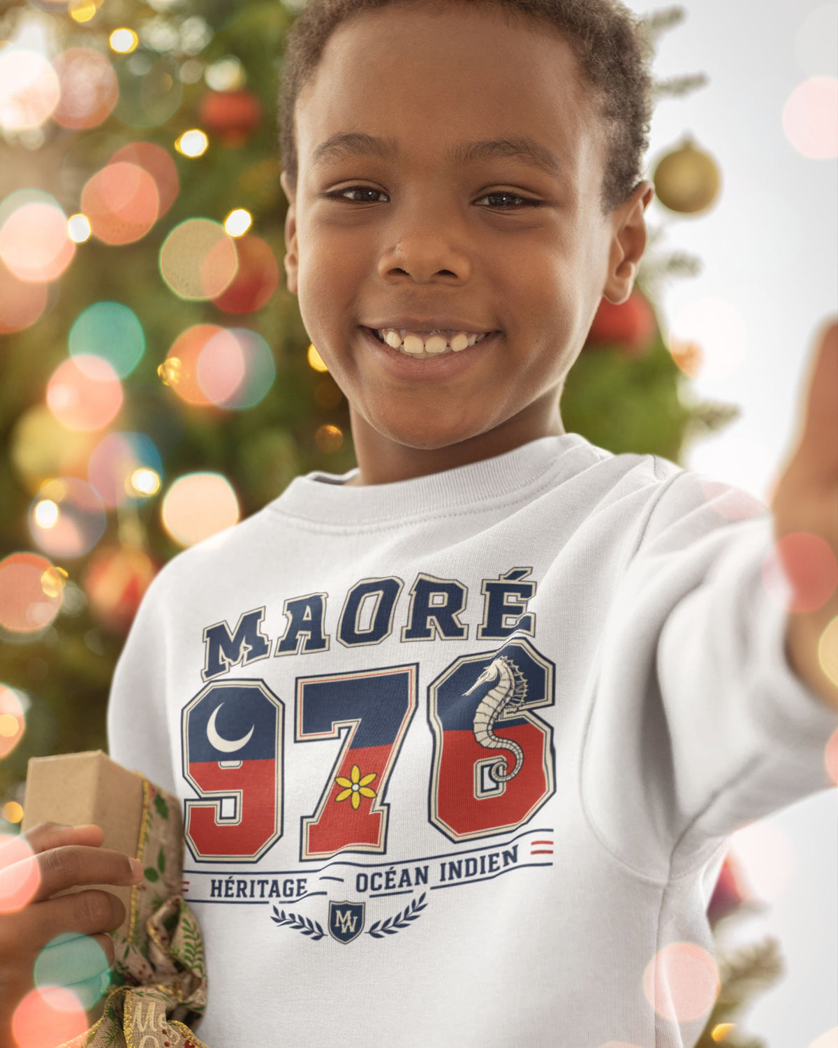  Enfant souriant portant le sweat blanc Maoré 976 Héritage, avec croissant de lune, fleur jaune et hippocampe, devant un sapin de Noël. 