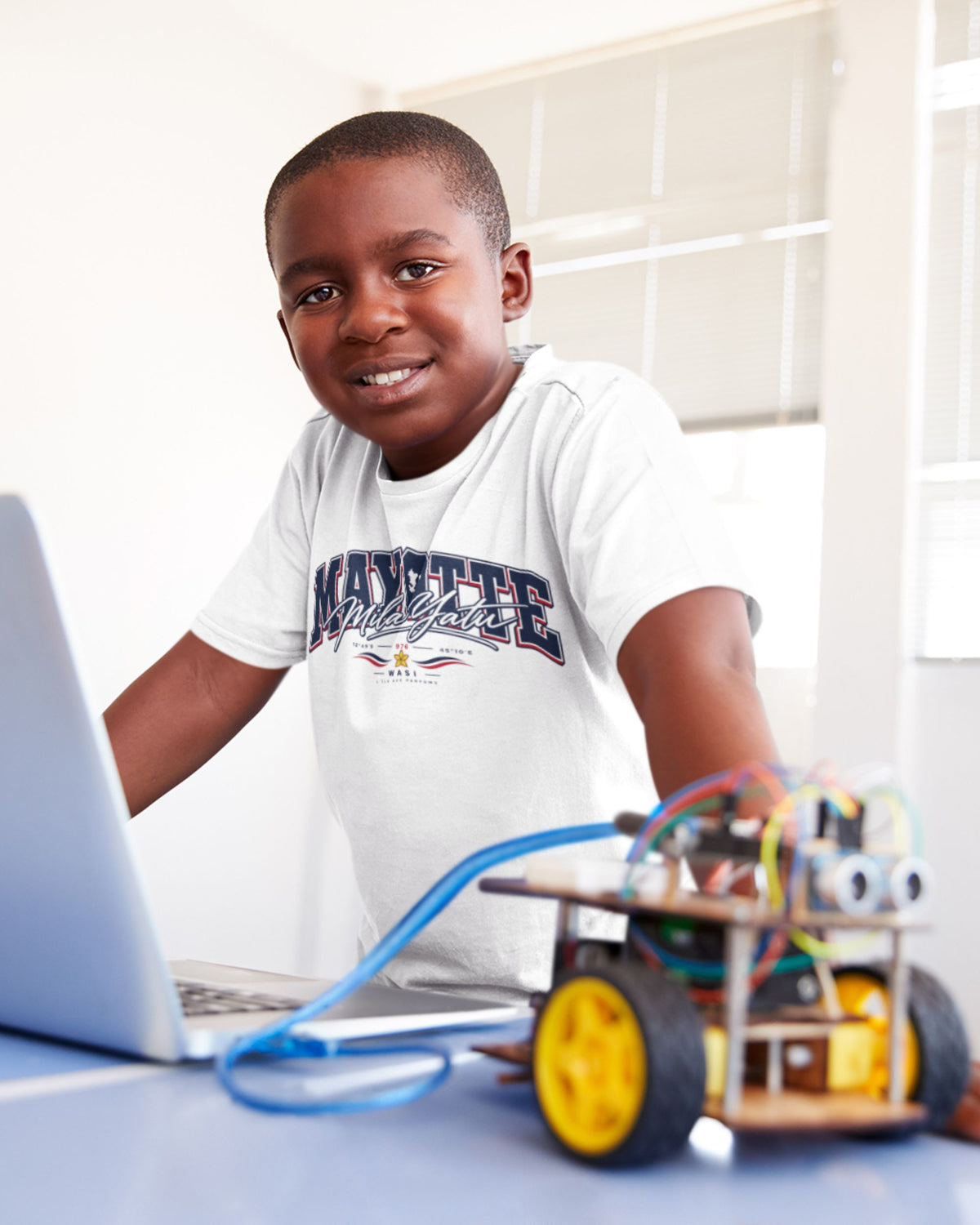 Enfant souriant portant le t-shirt blanc Mayotte Mila Yatu, devant un ordinateur portable et un petit robot à roues