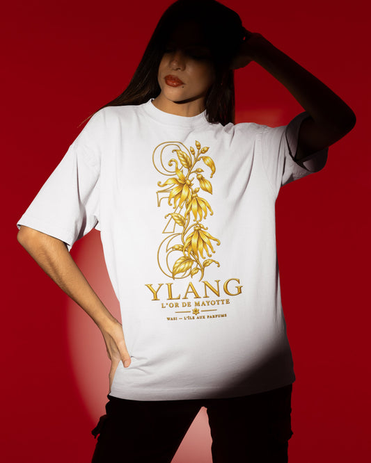 T-shirt oversize blanc WASI YLANG — L’Or de Mayotte, à motif d’ylang-ylang doré, porté par une femme sur fond rouge.