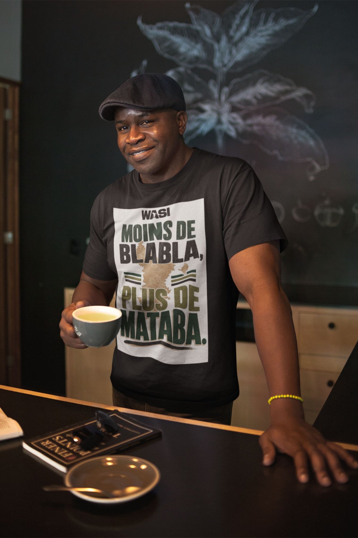 Homme tenant un café avec le t-shirt noir WASI Mataba, fond illustré ylang-ylang, ambiance lifestyle Maoré