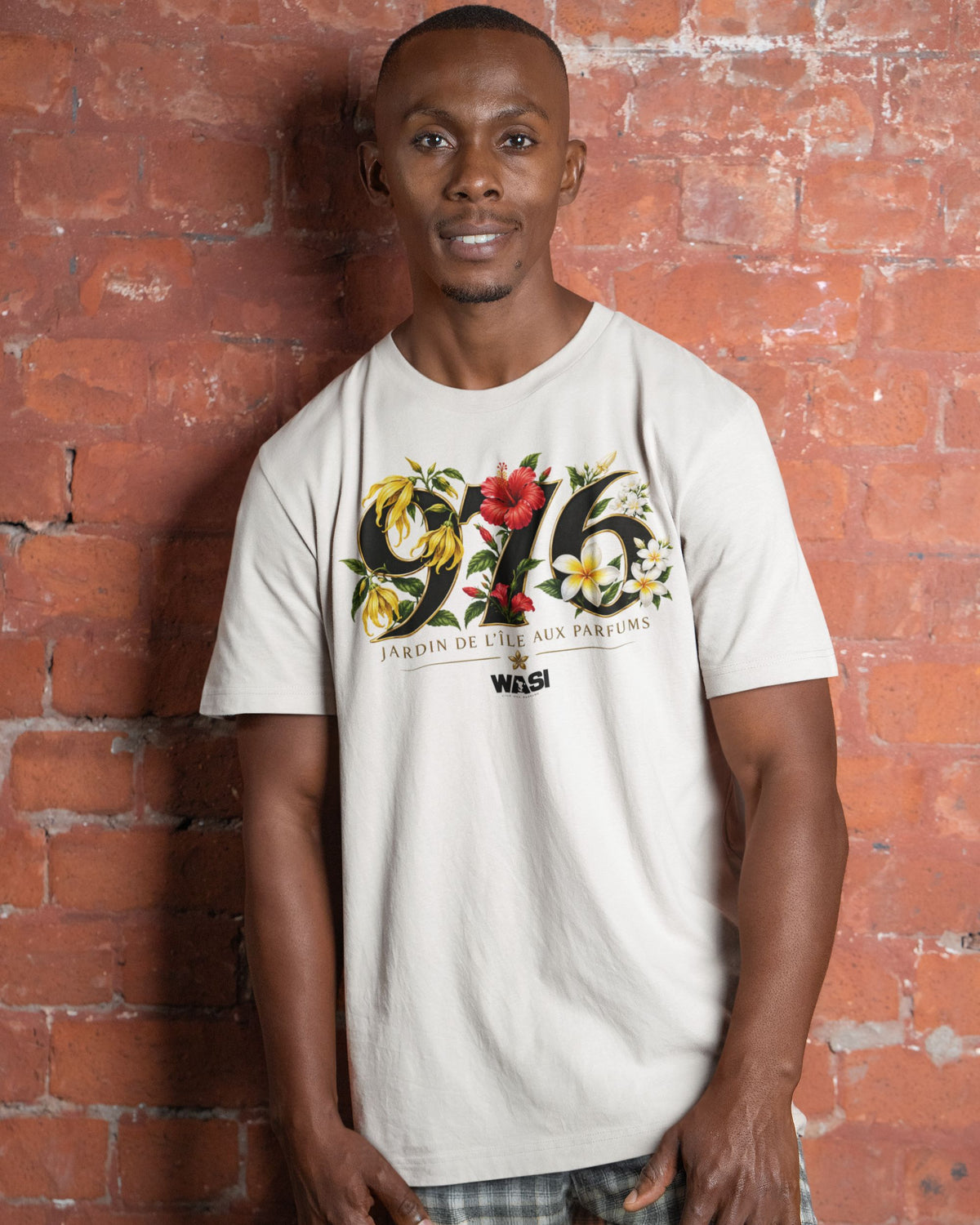 Homme portant le t-shirt ivoire WASI 976 Jardin de Maoré, inspiré des fleurs emblématiques de Mayotte, devant un mur en briques rouges.