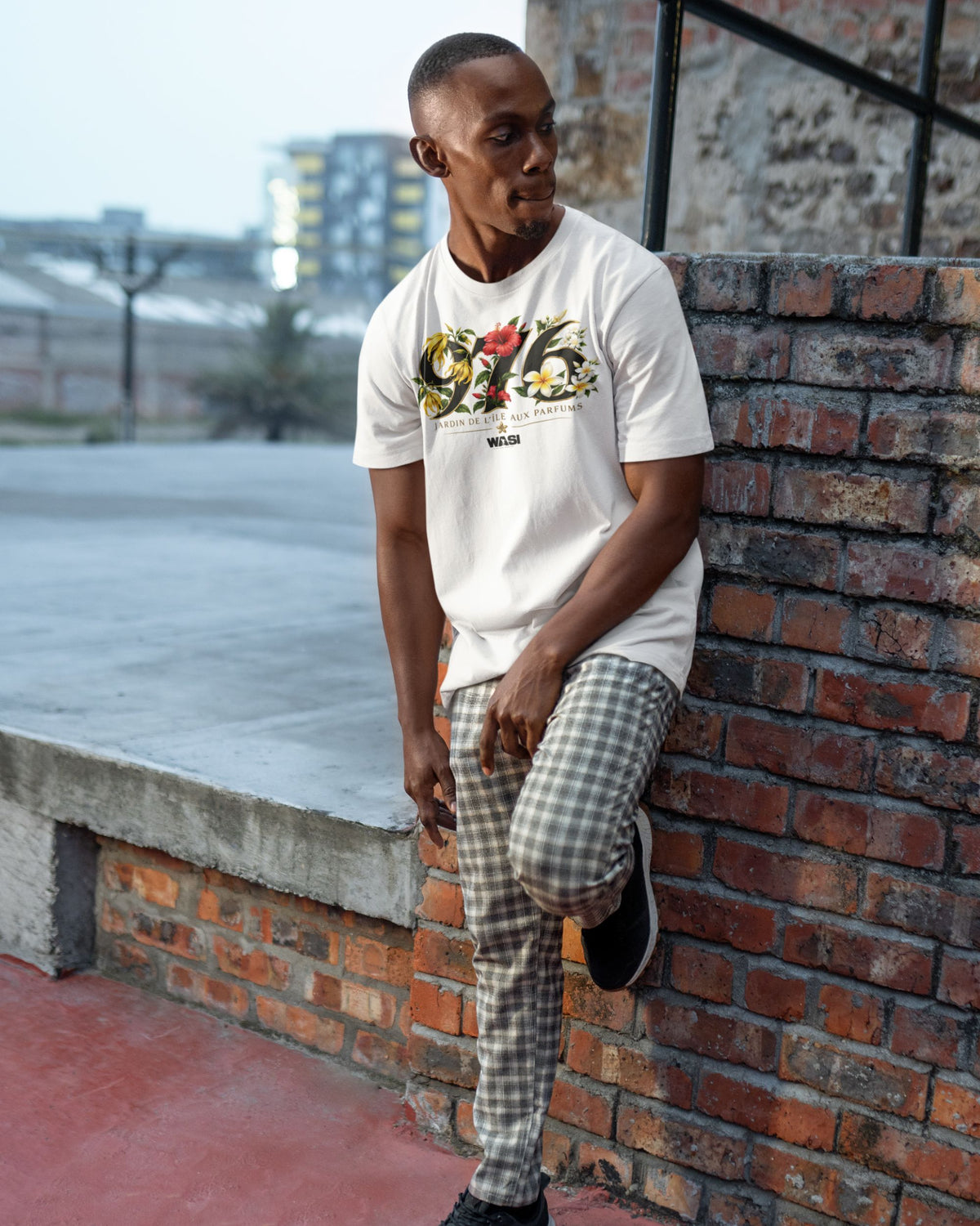 Homme portant le t-shirt ivoire WASI 976 Jardin de Maoré dans un décor urbain, adossé à un mur en briques, avec le design floral inspiré de Mayotte.