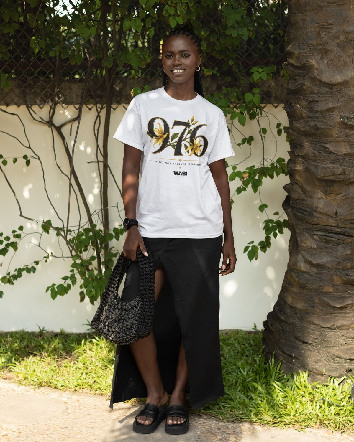 Femme portant un t-shirt blanc en coton WASI avec le design 976 et fleurs d'ylang-ylang, photographiée en extérieur dans un décor tropical inspiré de Mayotte.