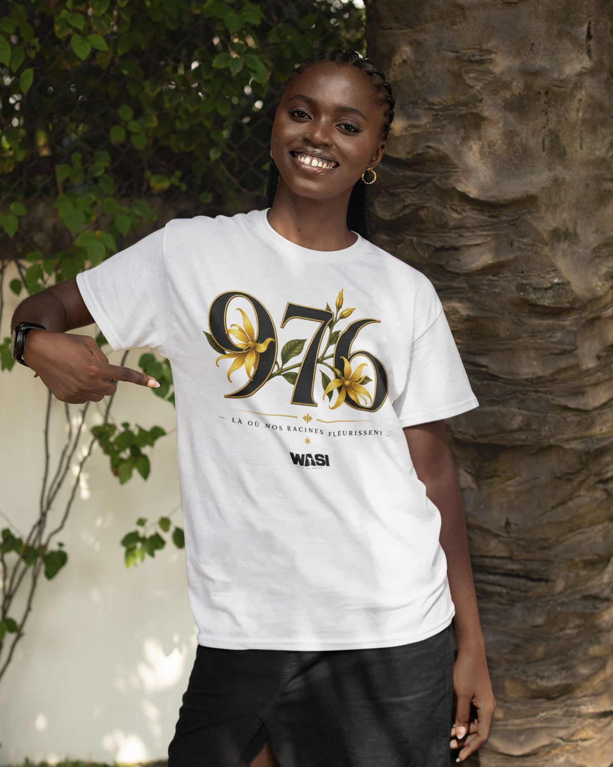 Femme souriante portant un t-shirt blanc WASI en coton avec le design 976 et fleurs d'ylang-ylang, pointant le motif dans un décor tropical inspiré de Mayotte.
