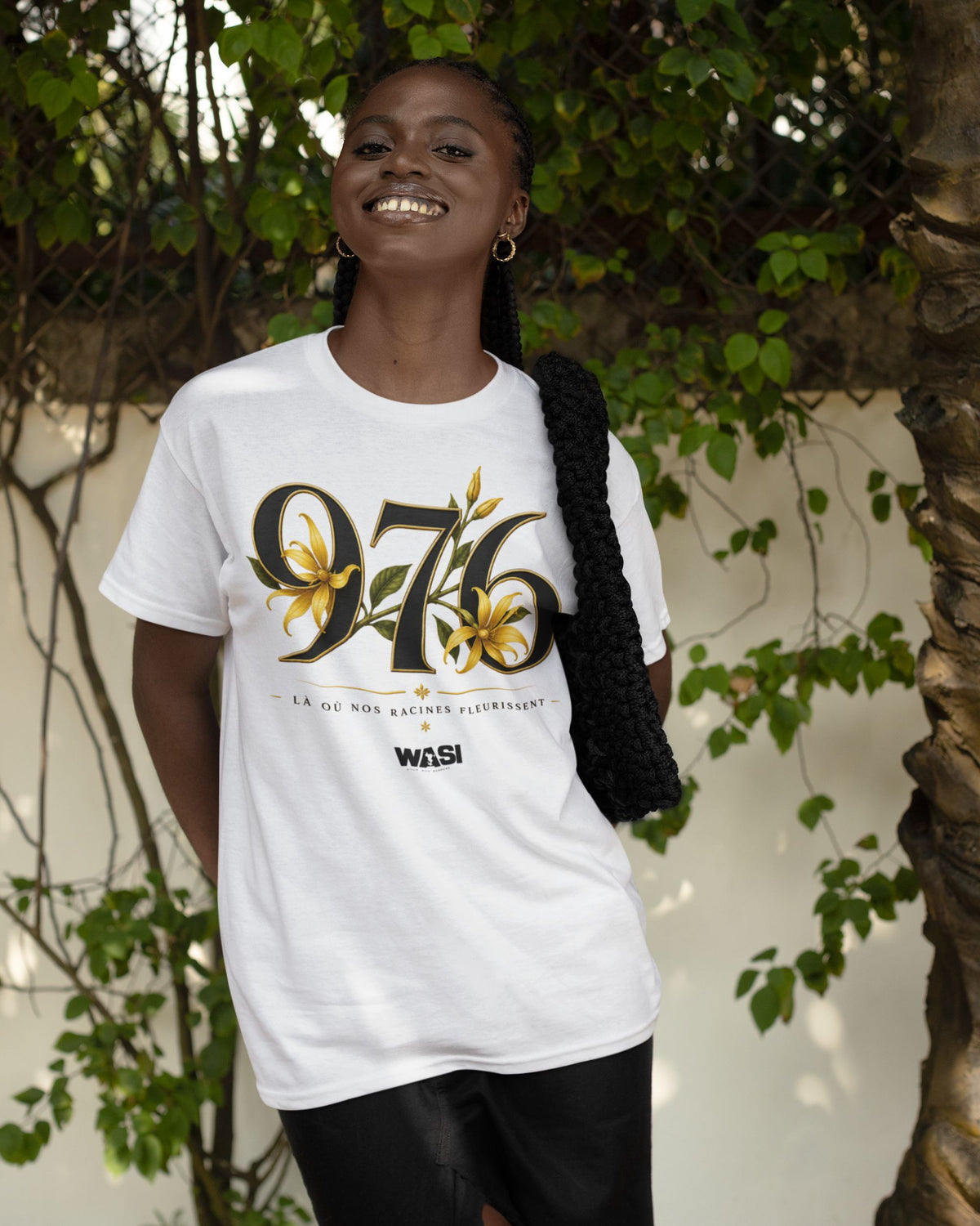 Femme portant un t-shirt blanc WASI avec le design 976 orné de fleurs d'ylang-ylang, photographiée en extérieur dans un décor végétal inspiré de Mayotte.